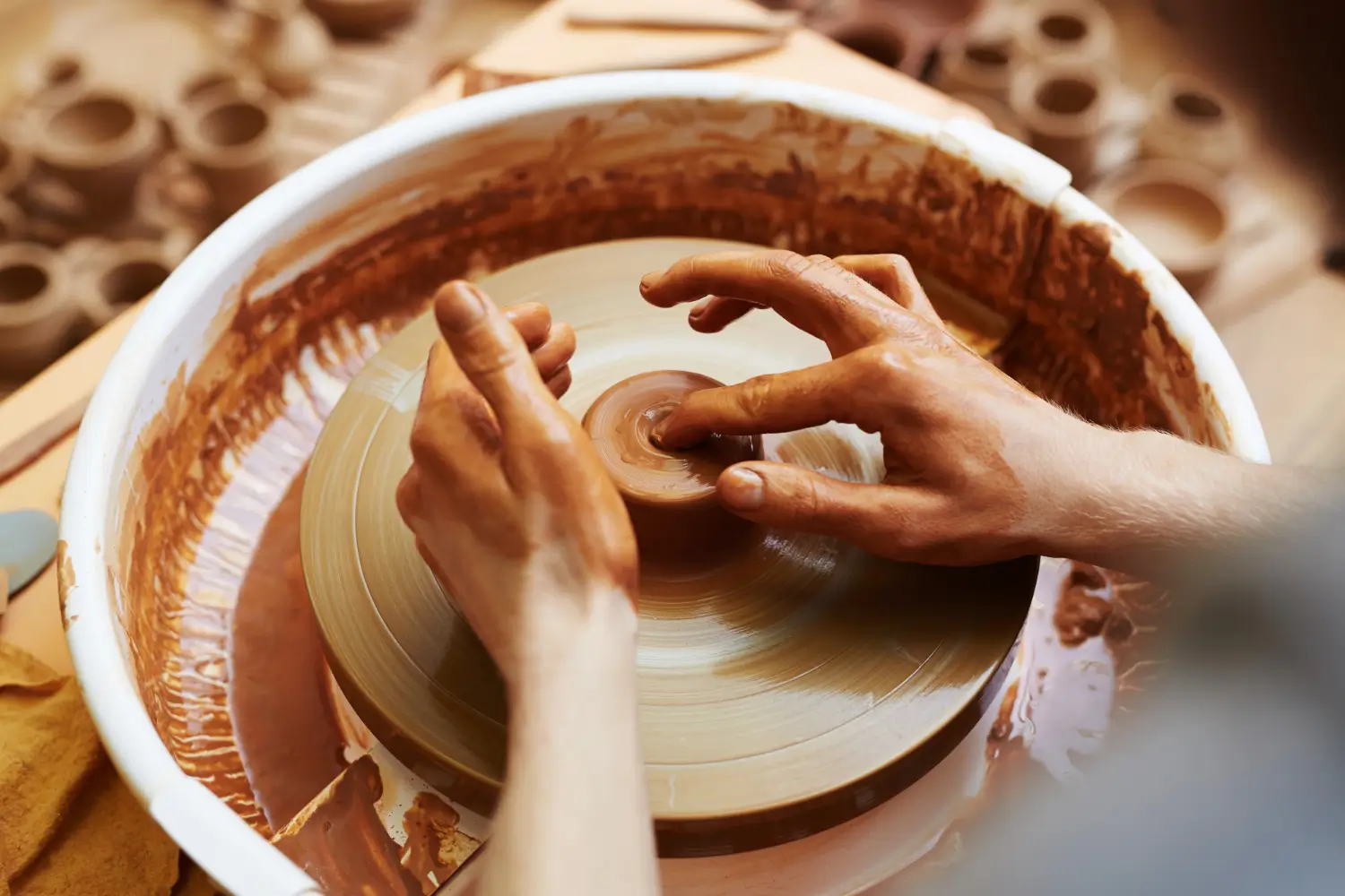 Manos moldeando arcilla en un torno de alfarero, creando una pieza de cerámica. El entorno muestra herramientas y productos de alfarería.
