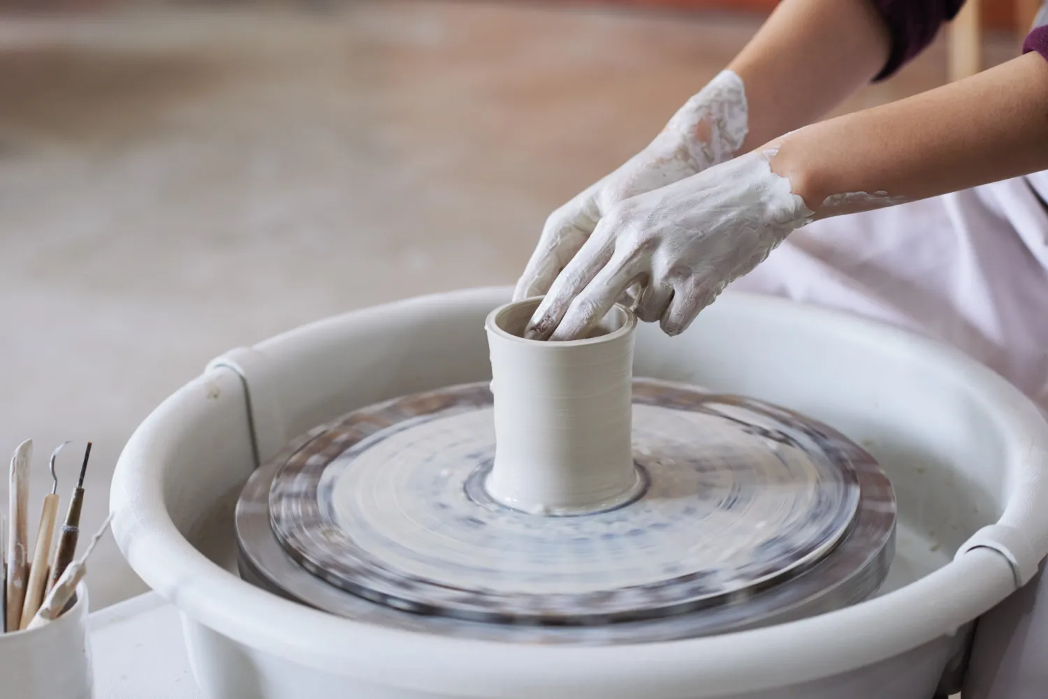 Manos de un alfarero moldeando una pieza de cerámica en un torno, con herramientas de alfarería a un lado y un ambiente artístico de fondo.