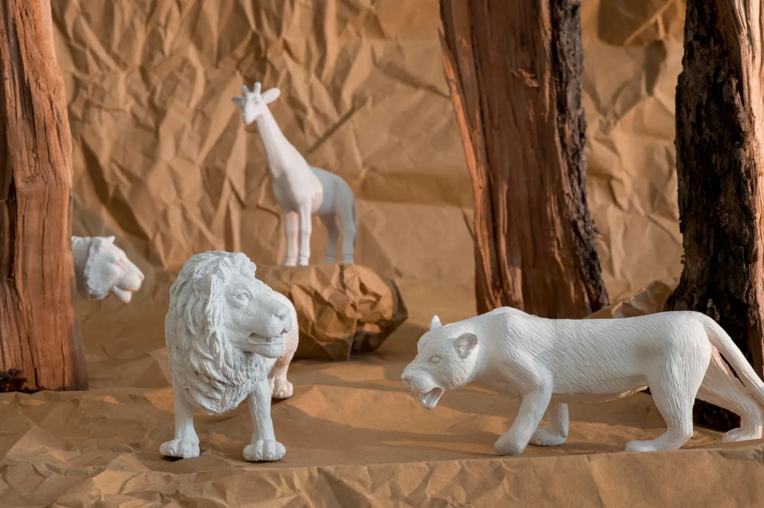 Figuras de animales en un entorno natural, incluyendo un león, un lobo y una jirafa, sobre un fondo de papel arrugado y troncos de madera.