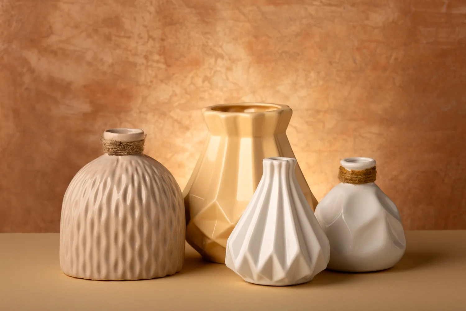 Vasos decorativos de cerámica en diferentes formas y colores, incluyendo tonos beige y blanco, sobre un fondo de textura cálida. Ideal para decoración de interiores.