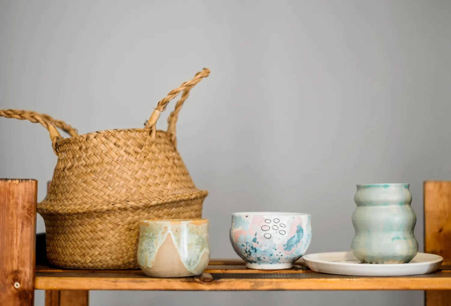 Conjunto de cerámica artesanal y una cesta de mimbre sobre una estantería de madera, presentando tazas y platos decorativos en tonos suaves y diseños únicos.