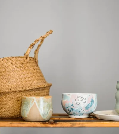 Conjunto de cerámica artesanal y una cesta de mimbre sobre una estantería de madera, presentando tazas y platos decorativos en tonos suaves y diseños únicos.
