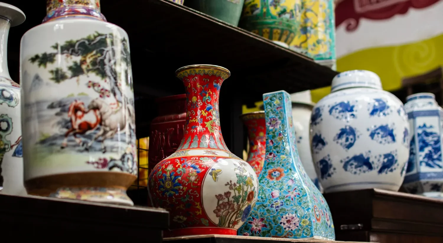 Vasos de cerámica decorativos de diferentes colores y estilos, exhibidos en una estantería. Los diseños incluyen motivos florales y escenas de la naturaleza, destacando la artesanía tradicional.